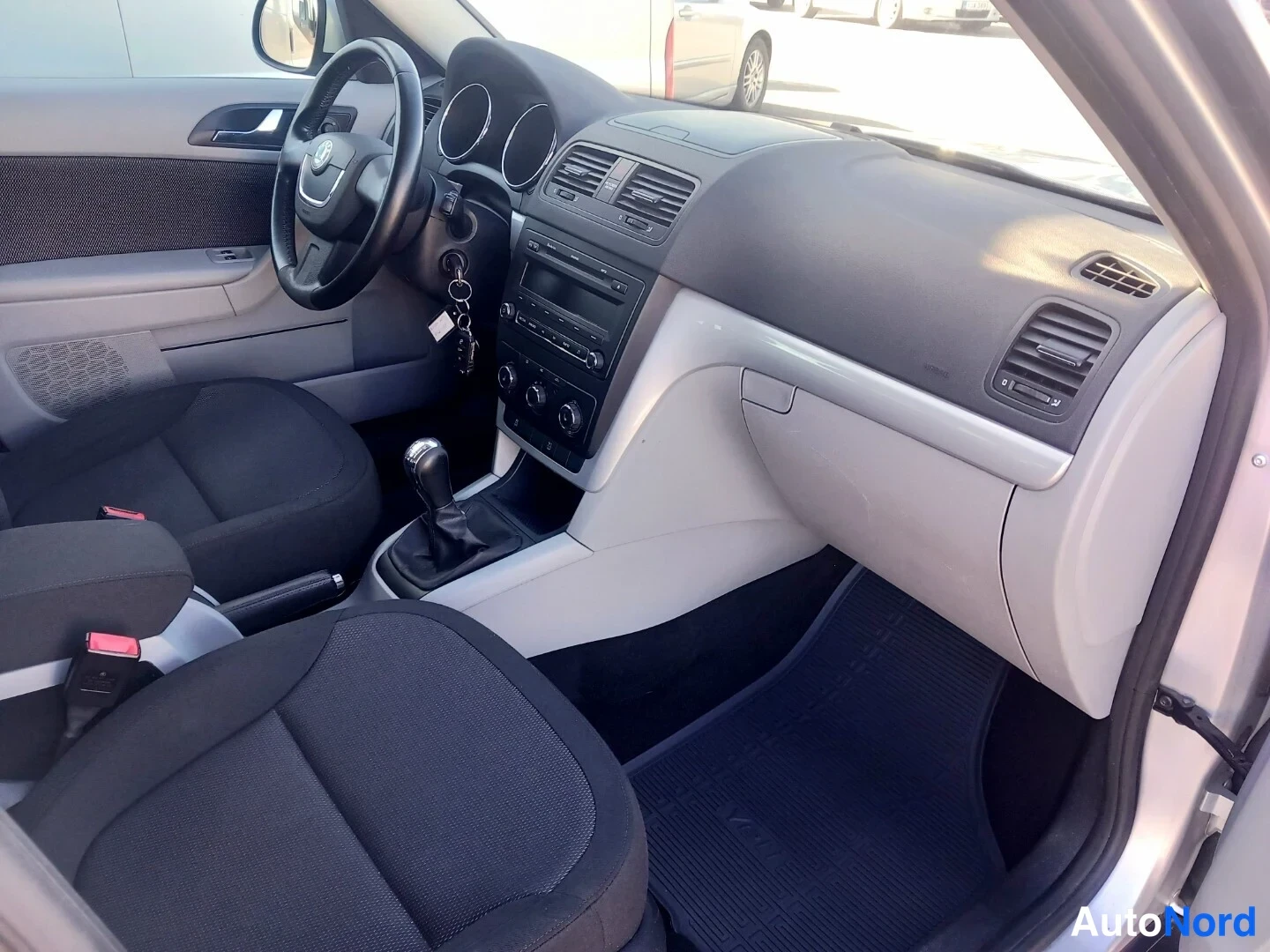 Etuistuimet: Interior view of the front cabin showing black fabric driver and passenger seats, a manual gear lever in the centre console, a dark dashboard, and an open glove