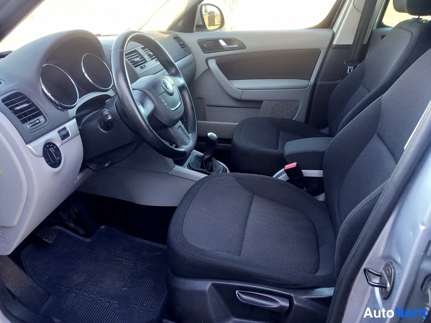 Etuistuimet: Front cabin interior with two black fabric seats, a multi-function steering wheel, instrument cluster, manual gear stick, and door handle on the left side.