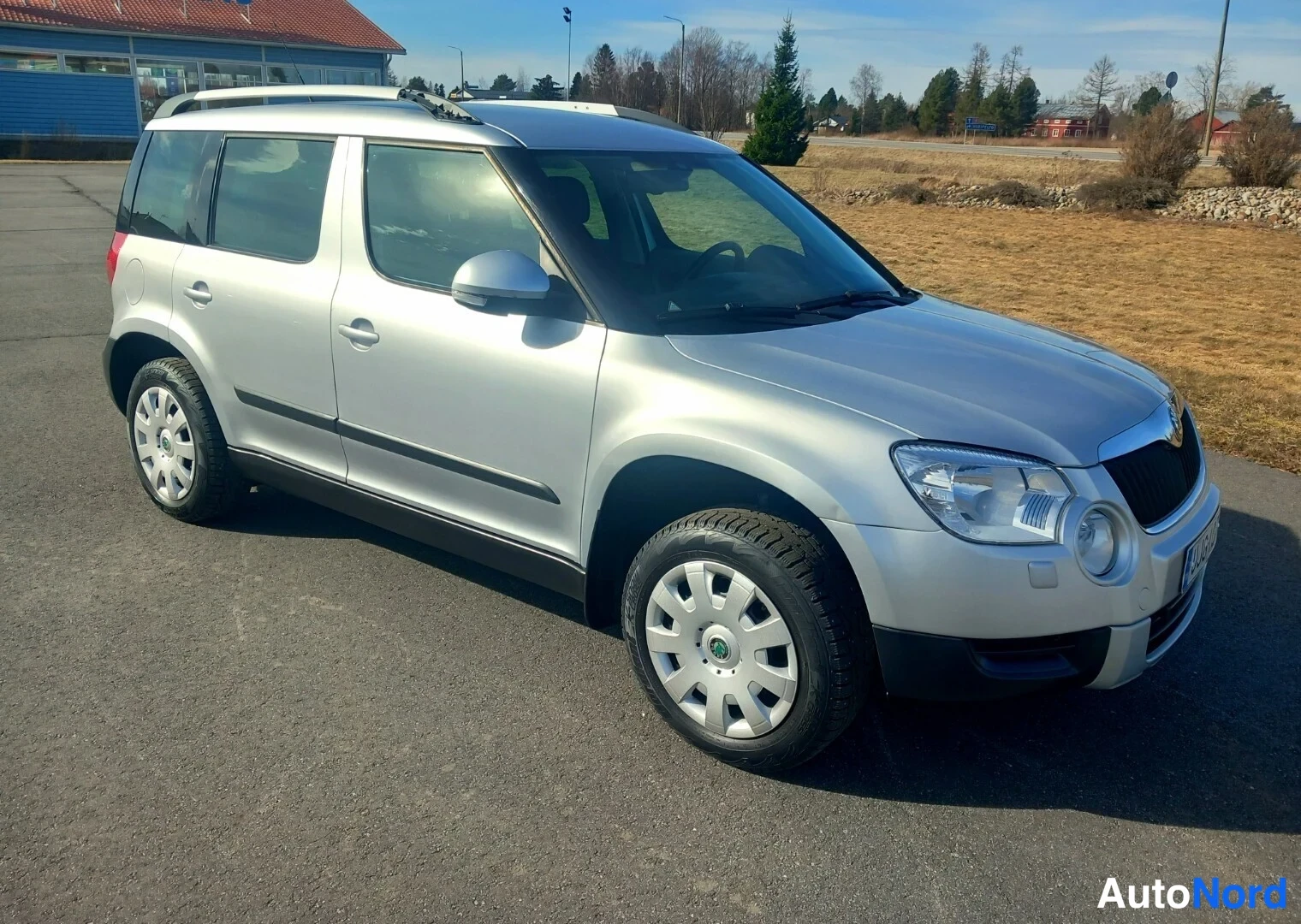 Etuviisto, nokka oikealle: Silver Skoda Yeti photographed at a front three-quarter angle on asphalt with roof rails visible; grille and headlights appear in clear halogen form unde