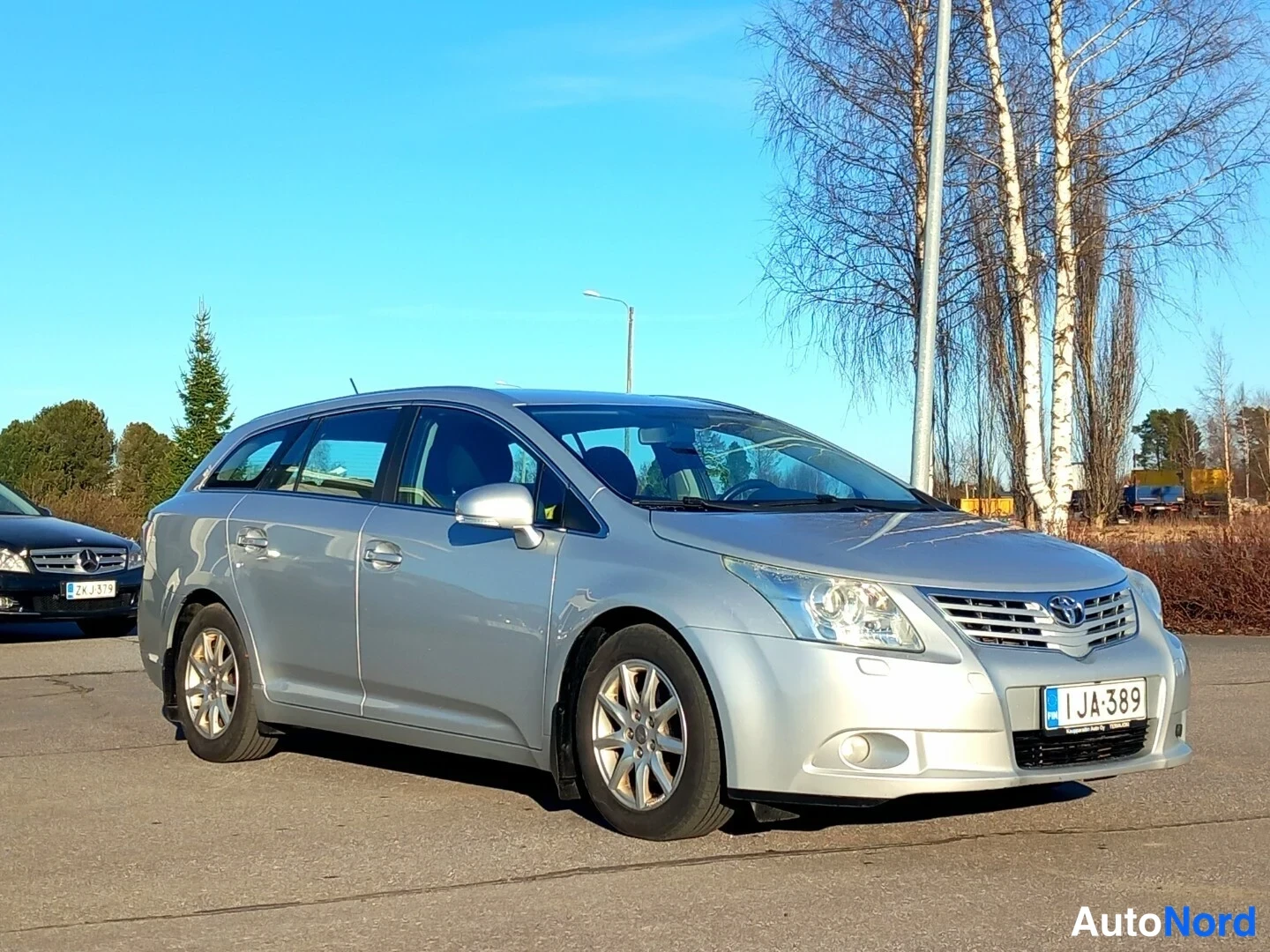 Etuviisto, nokka vasemmalle: Toyota Avensis -farmari edestä vasemmalta sivulta aurinkoisena päivänä, hopeisenä, rekisteritunnus.