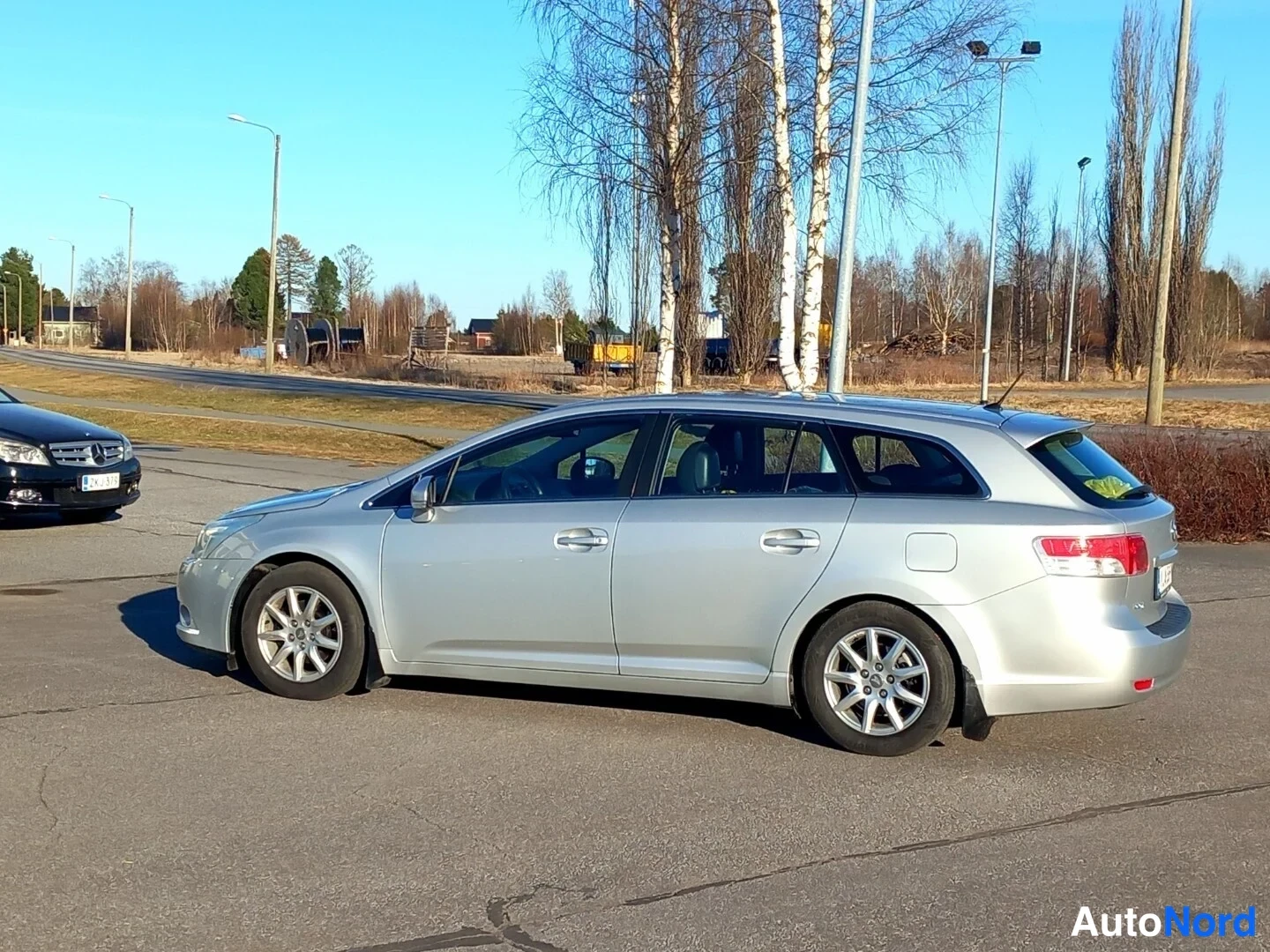 Takaviisto: Hopeanhohtoinen farmariauto takaosalta ja sivulta, perä oikealla, auringonpaisteessa parkkialueella.