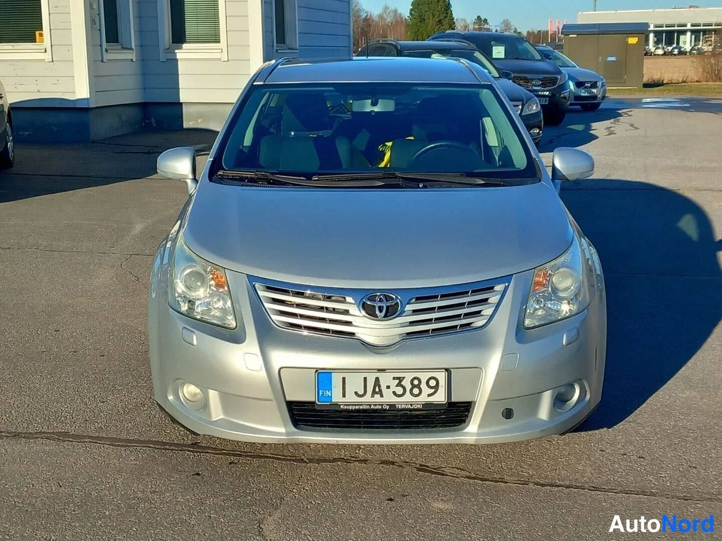 Edestä: Toyota Avensis -farmarin etupää edestä kuvattuna, rekisterikilpi, hopeakorinen auto.