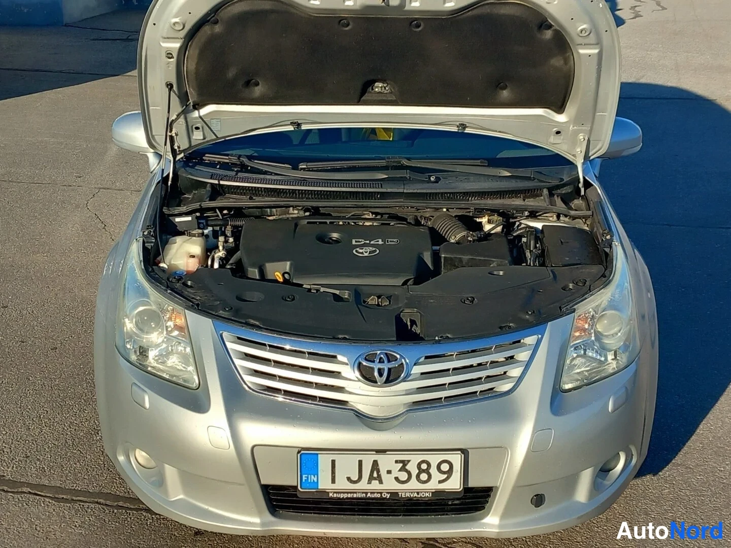 Edestä: Toyota Avensisin konepelti auki, D-4D-merkitty moottoritila ja Toyota-merkintä näkyvissä, etupää suoraan edestä.