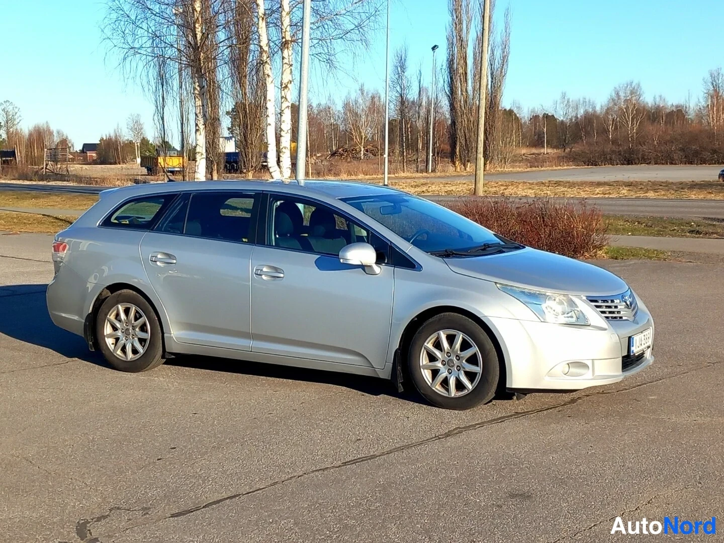 Etuviisto, nokka oikealle: Hopeanvärinen Toyota Avensis -farmari kolmasosakulmasta keula oikealla, koko auto näkyvissä parkkipaikalla.