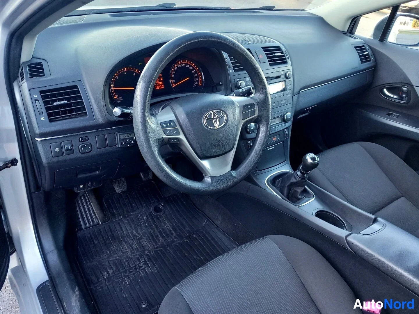 Edestä: Auton ohjaamo vasemmalta: Toyota-ohjauspyörä, analogiset mittarit, manuaalivaihteisto ja tummat kangasistuimet.
