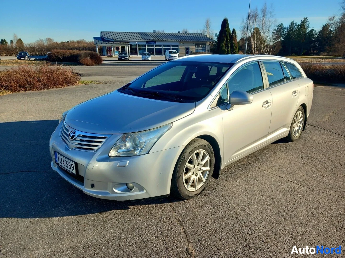 Etuviisto, nokka vasemmalle: Hopeinen Toyota Avensis -farmari edestä vasemmalla sivulla parkkipaikalla, rekisteritunnus.