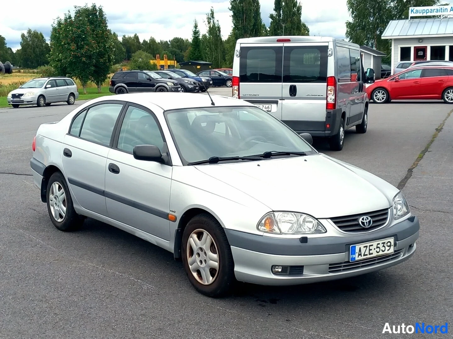 Etuviisto, nokka oikealle: Toyota Avensis -sedanin etukulma toisesta suunnasta: neliovinen hopeakarinen korirakenne, rekisterikilpi.