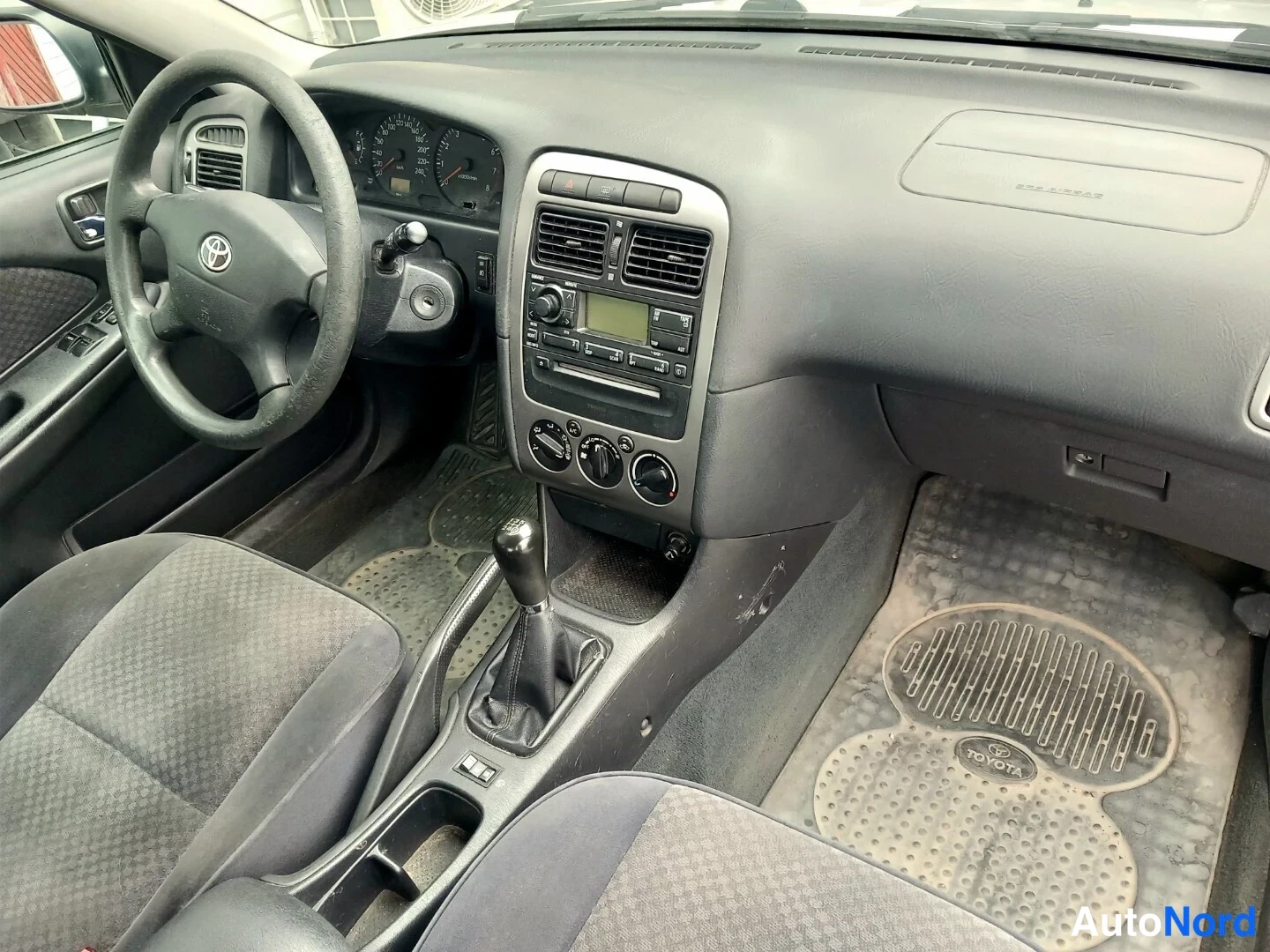 Muu: Sisätilan yleiskuva kojelaudasta: Toyota-ohjauspyörä, manuaalivaihteisto, radiopanelo ja Toyota-merkkiset mattopyyheet.