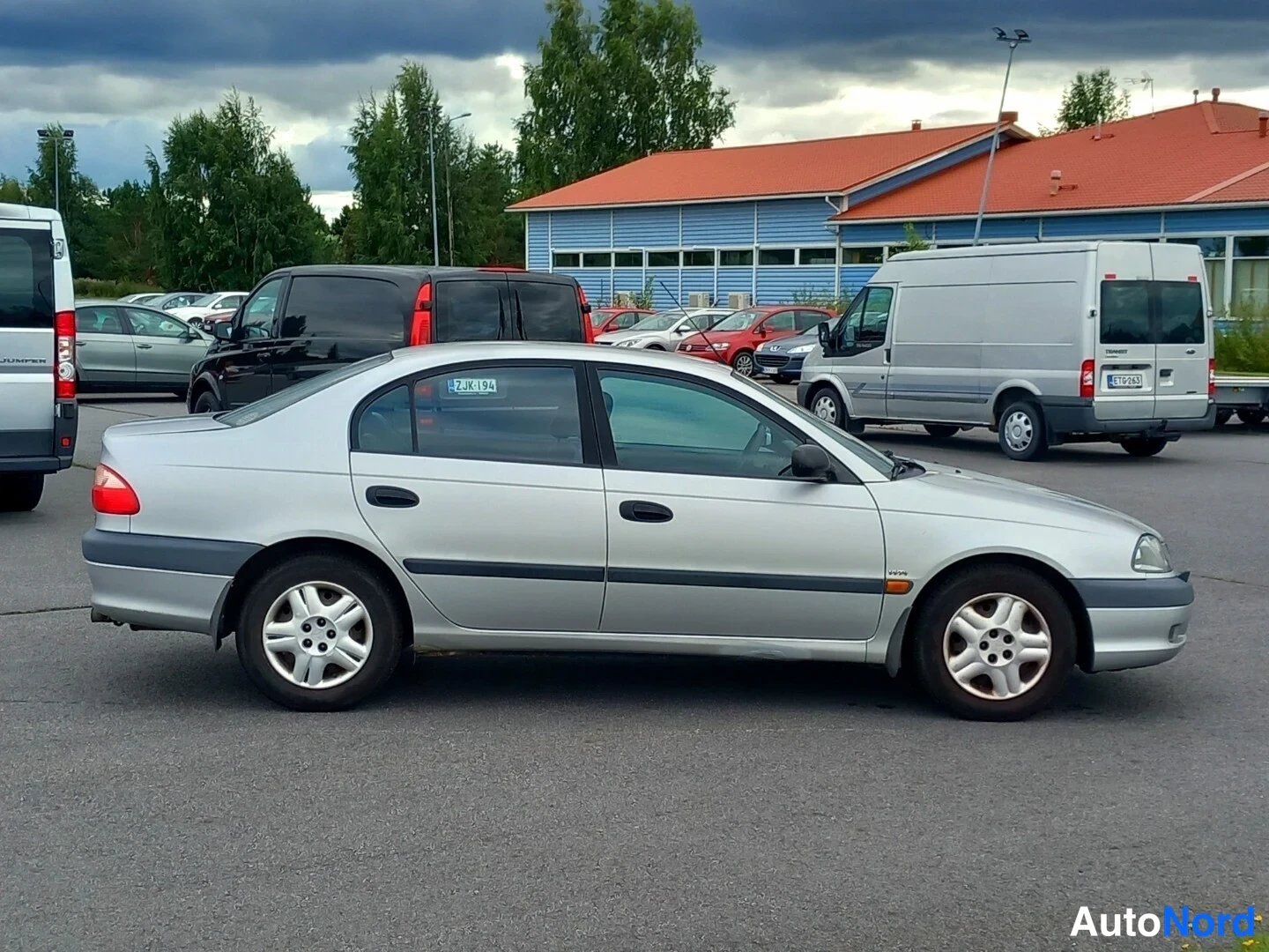 Takaviisto: Hopeanvärinen sedan henkilöauto sivulta-takaapäin kuvattuna parkkipaikalla.