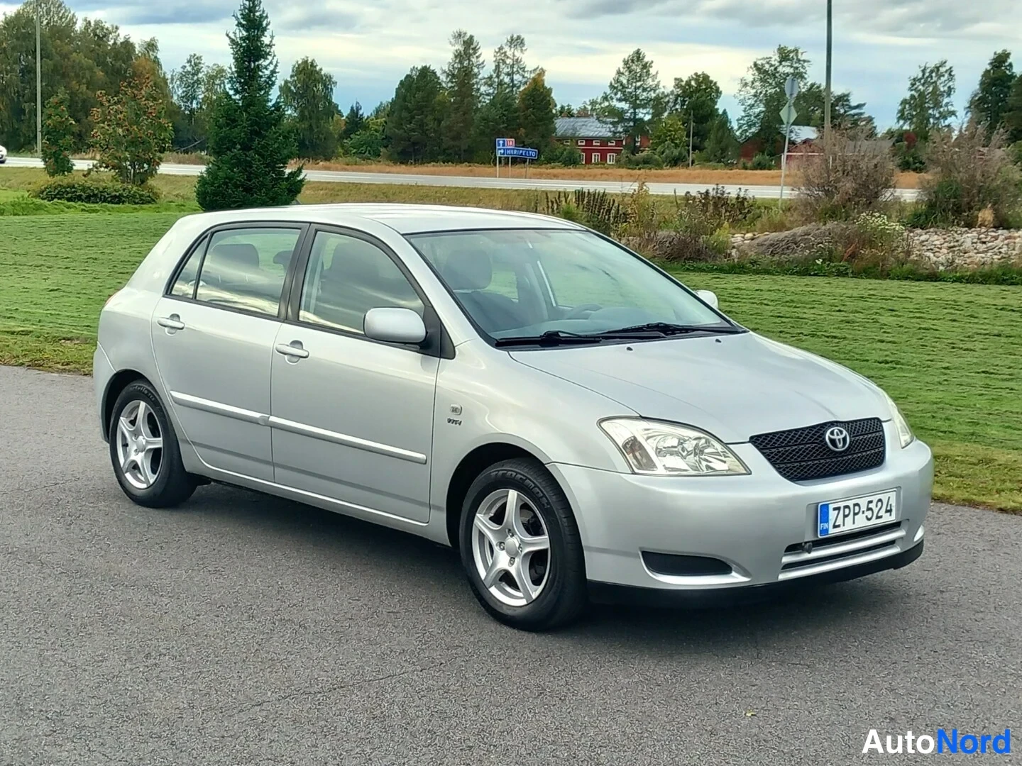 Etuviisto, nokka oikealle: Hopeanvärinen Toyota Corolla viistosti edestä oikealta, rekisteritunnus, takana nurmikenttä ja tie.