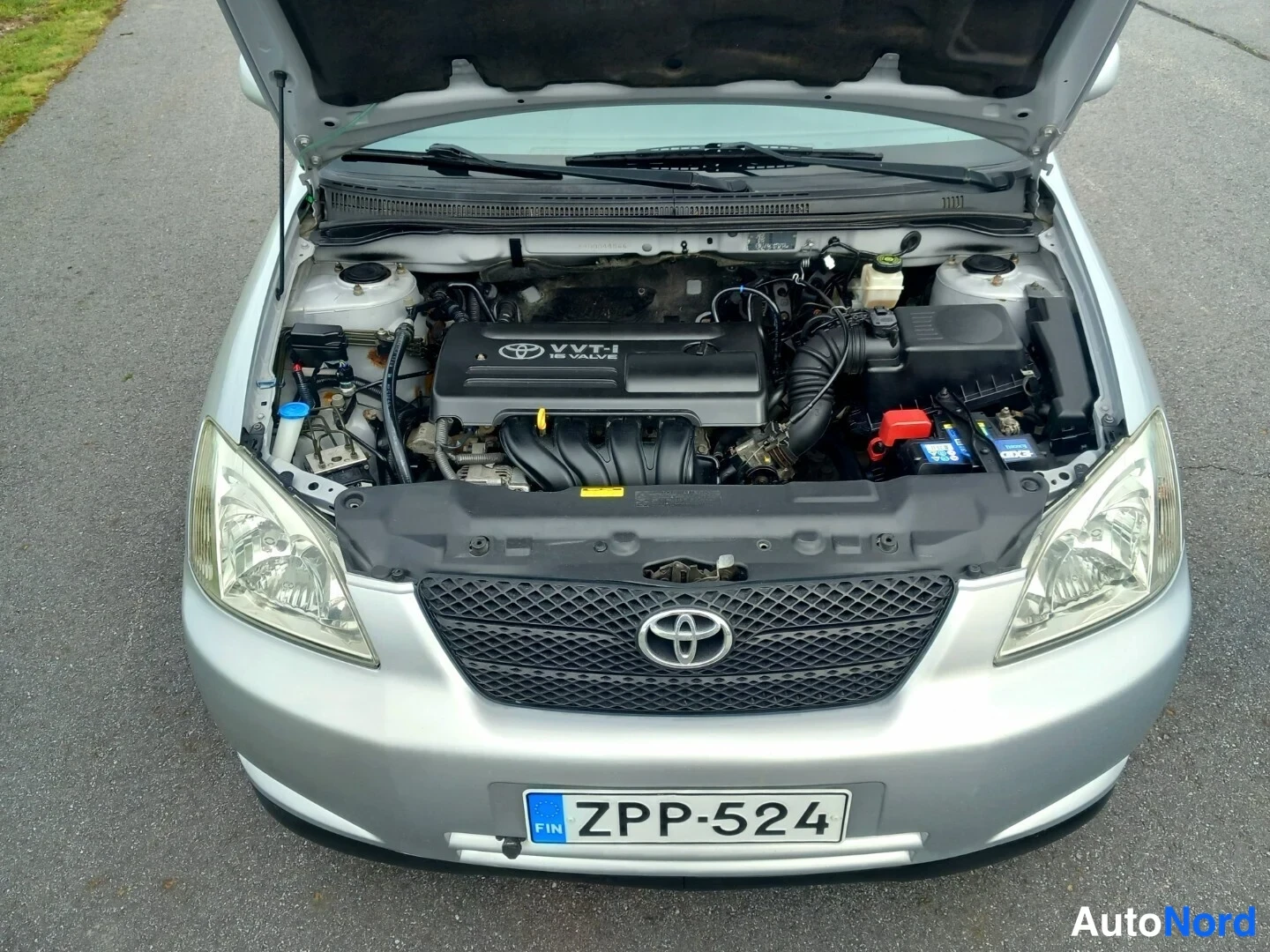 Edestä: Konepelti auki hopeanvärisessä Toyota Corollassa; VVT-i -merkitty moottori ja akku näkyvät konepellin alla.