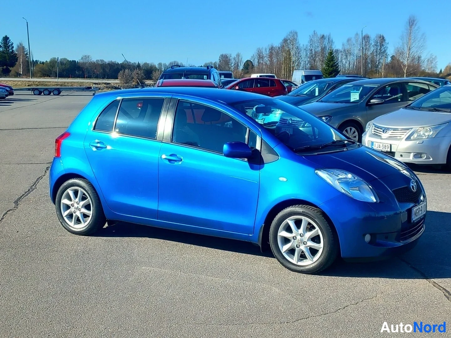 Etuviisto, nokka oikealle: Sininen Toyota Yaris -henkilöauto kuvattu kulmasta edestä oikealta, etusäleikkö ja ajovalot näkyvät, hopeavanteet.