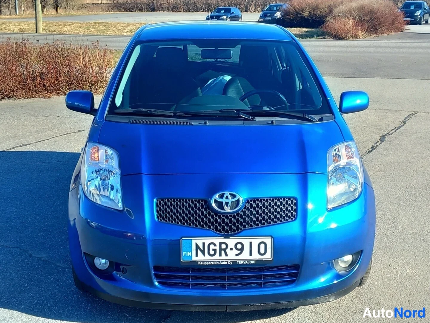 Edestä: Sininen Toyota Yaris kuvattuna suoraan edestä: Toyota-merkki mustassa verkkogrillissä, sumuvalot puskurissa ja rekisteritunnus.