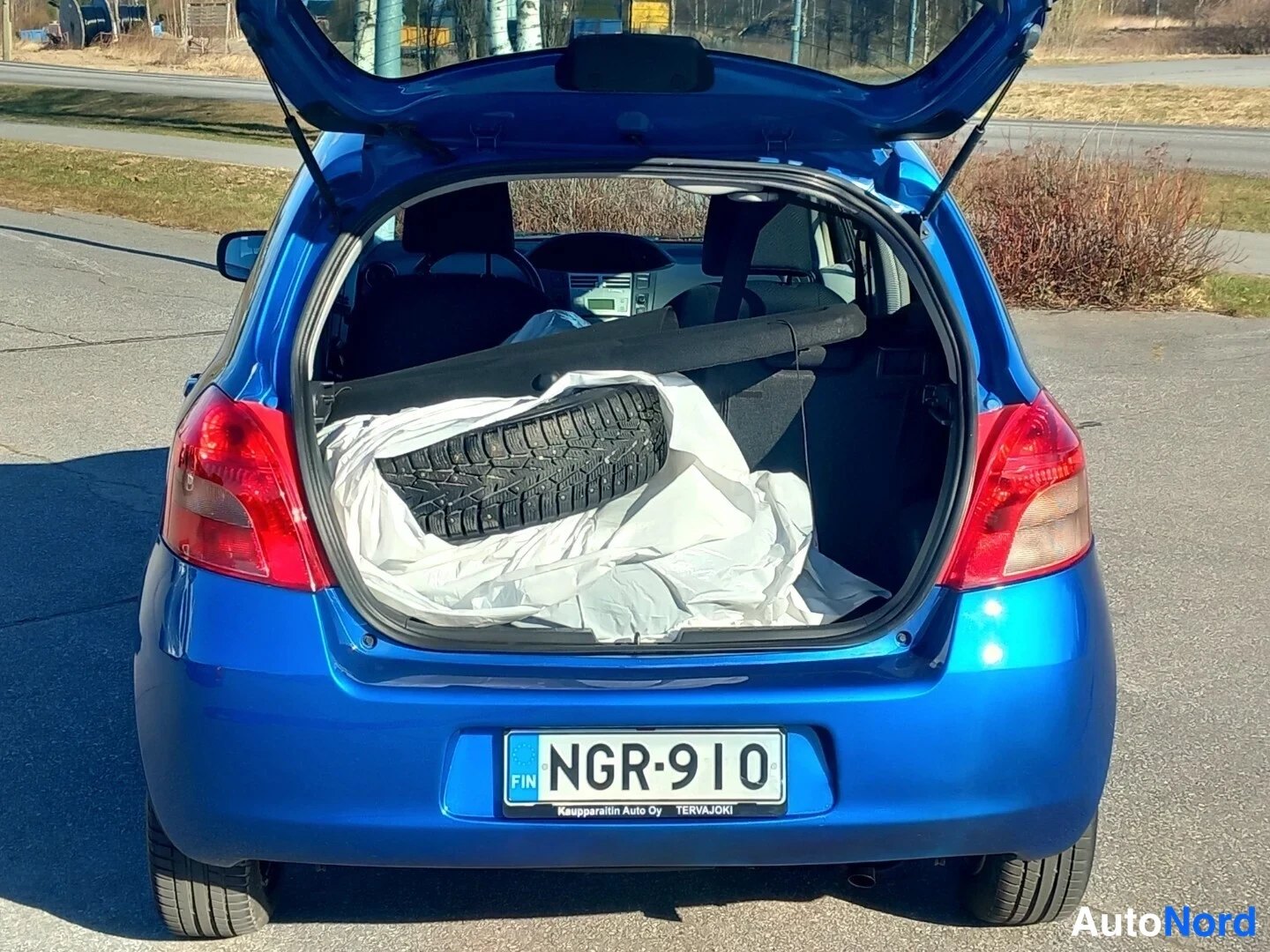 Takaa: Sinisen auton tavaratila auki takaa kuvattuna: tavaratilassa niitattu talvirengas valkoisessa muovipussissa ja irti nostettu tavaratilan suoja, rekisterikilpi.