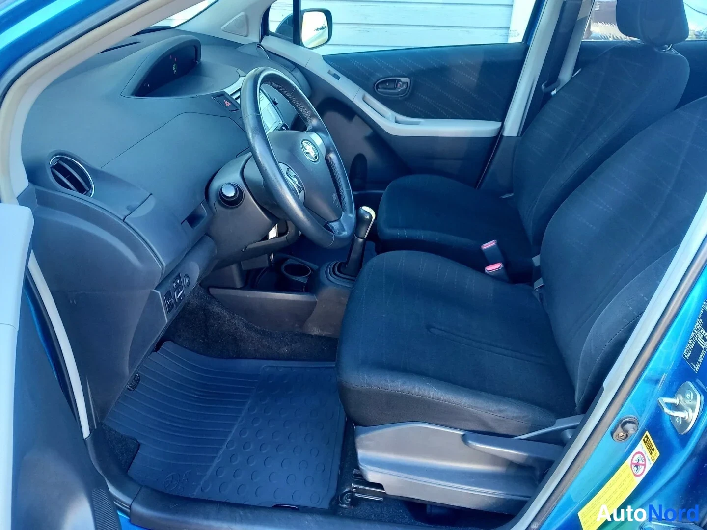 Muu: Kuljettajan puolen näkymä Toyota Yarikseen: ohjauspyörä, manuaalivaihde, mustat kangastuolit ja kumimatto.