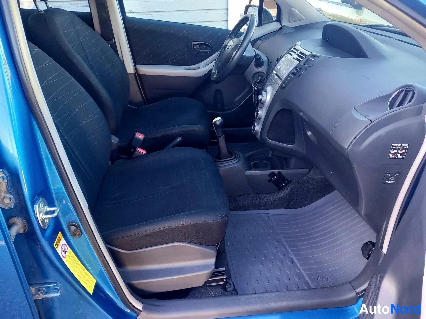 Muu: Avatusta etuovesta kuvattu Toyota Yarisin sisätila: kaksi etuistuintä, manuaalivaihteisto ja kumimatot.