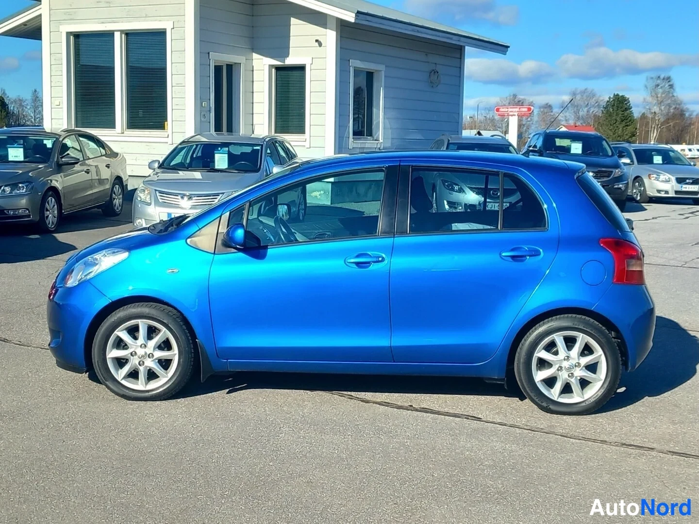 Etuviisto, nokka vasemmalle: Sininen Toyota Yaris -henkilöauto sivulta etukulmasta aurinkoisella parkkipaikalla.