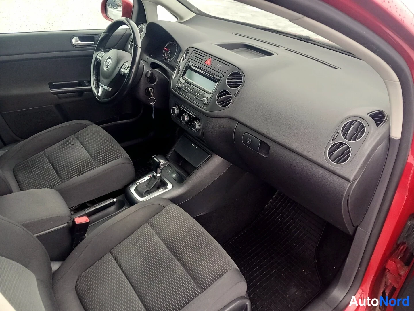 Muu: Autoa kuvattu avoimesta etuovesta: tumma sisusta, Ohjaamo, kojelauta ja etupenkit nakyvat. Volkswagen-ohjauspyora, automaattivaihteiston valitsin, radio ja ilma