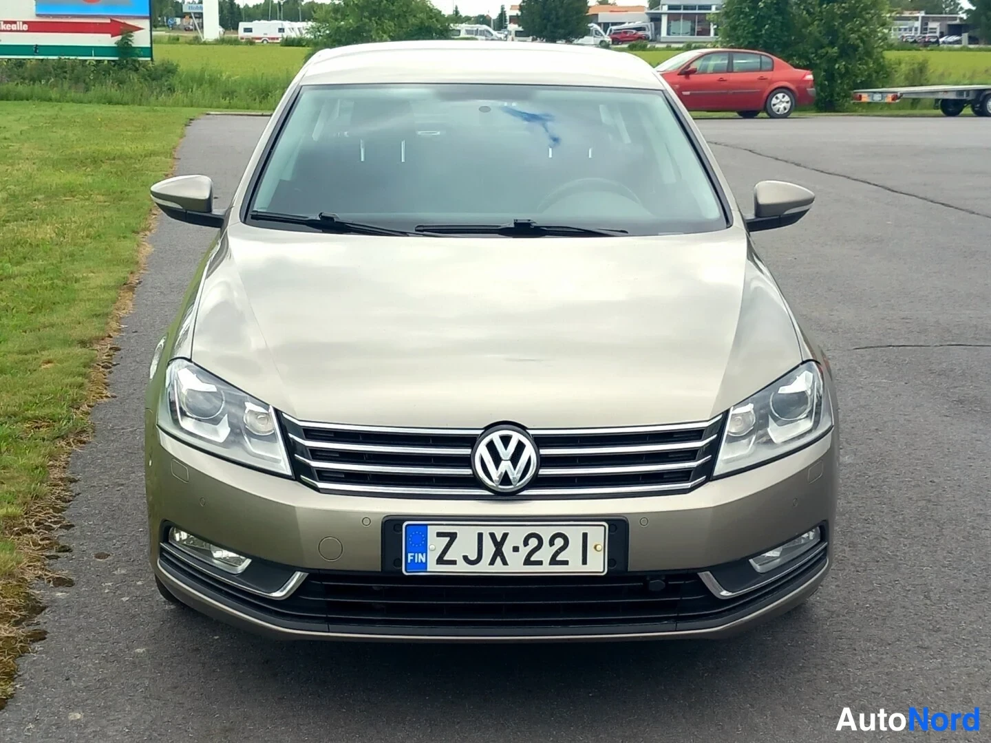 Edestä: Volkswagen Passatin suora etukuva: kultaharmaa keula, VW-merkki, rekisterikilpi, asfalttiparkkipaikalla ja nurmikko vasemmalla.
