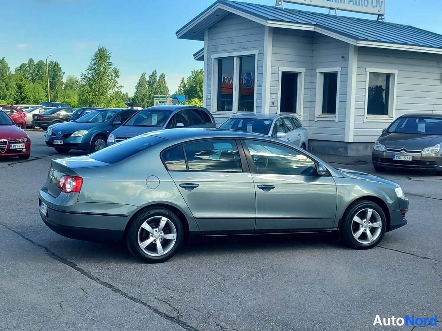 Takaviisto: Volkswagen Passat -sedan takaosalta oikealta sivulta, harmaa metallinen, autopalvelun rakennus taustalla.