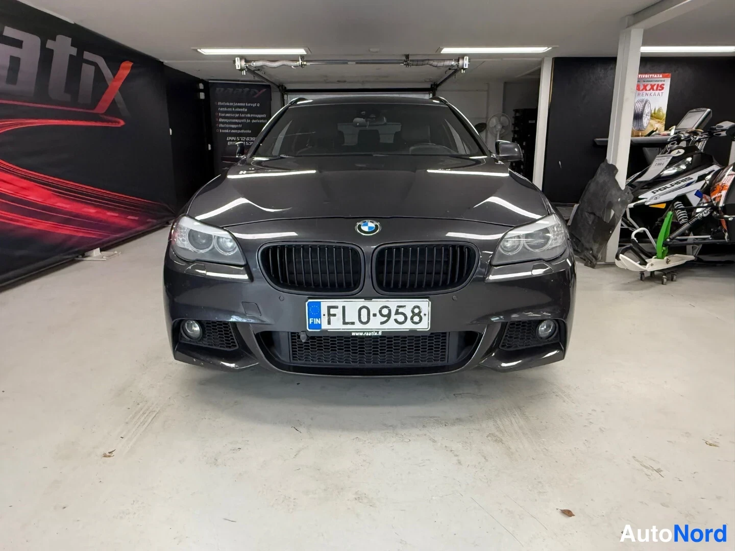 Edestä: BMW:n etupää suoraan edestä kuvattuna tummana autona hallissa, rekisterikilpi FL0-958.