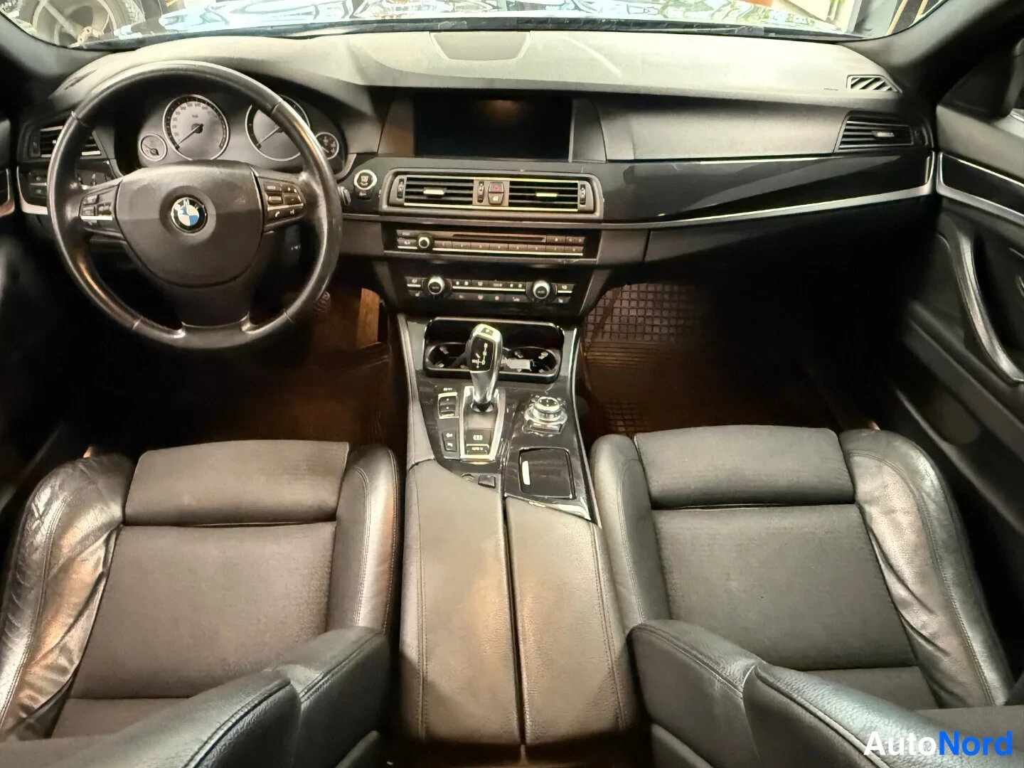 Yksityiskohta: Auton sisätila: ohjaamo, jossa BMW-ohjauspyörä, kojelauta, keskikonsoli automaattivaihteen valitsimella ja kaksi mustaa penkkiä.