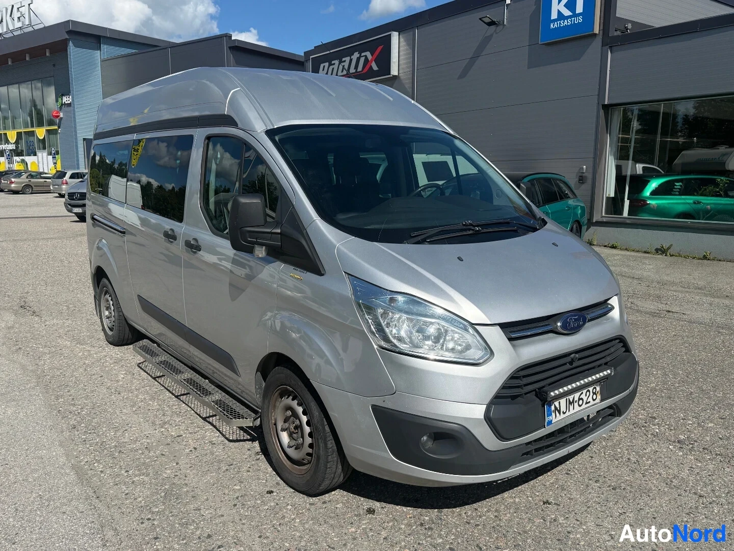 Etuviisto, nokka oikealle: Hopeanvärinen Ford Transit Custom -pakettiauto etuviistöstä oikealta, korkeakattoinen ja tummennetuilla ikkunoilla, rekisterikilpi.