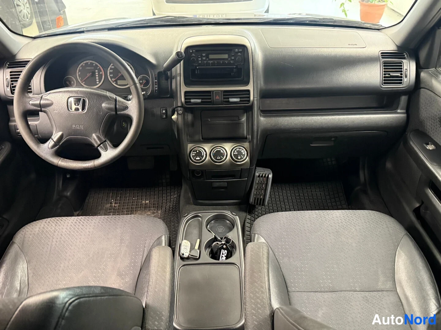 Muu: Honda CR-V:n ohjaamosta edestä: Honda-ohjauspyörä, mittaristo, keskikonsolin radio ja kolme pyöreää säädintä, kangasistuimet ja jalkamatot.