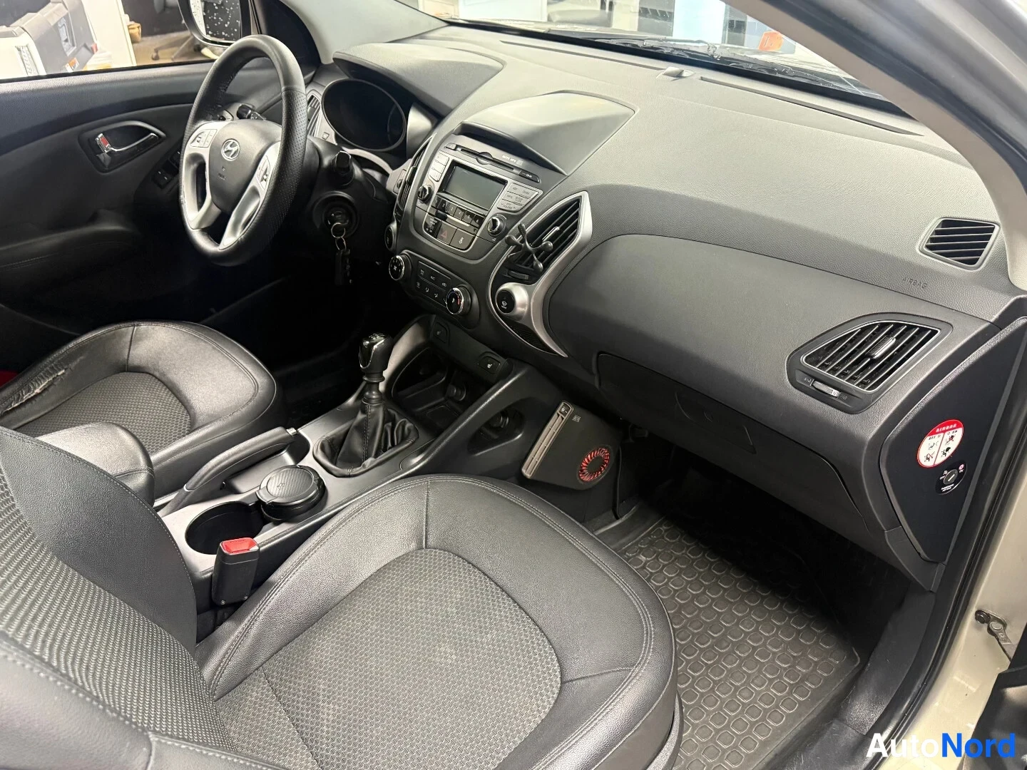 Muu: Hyundai ix35:n ohjaamo kuvattuna matkustajan puolelta: musta kojelauta, manuaalivaihteen vaihtopyörä, tummat istuimet ja kuviolliset lattiamatot.