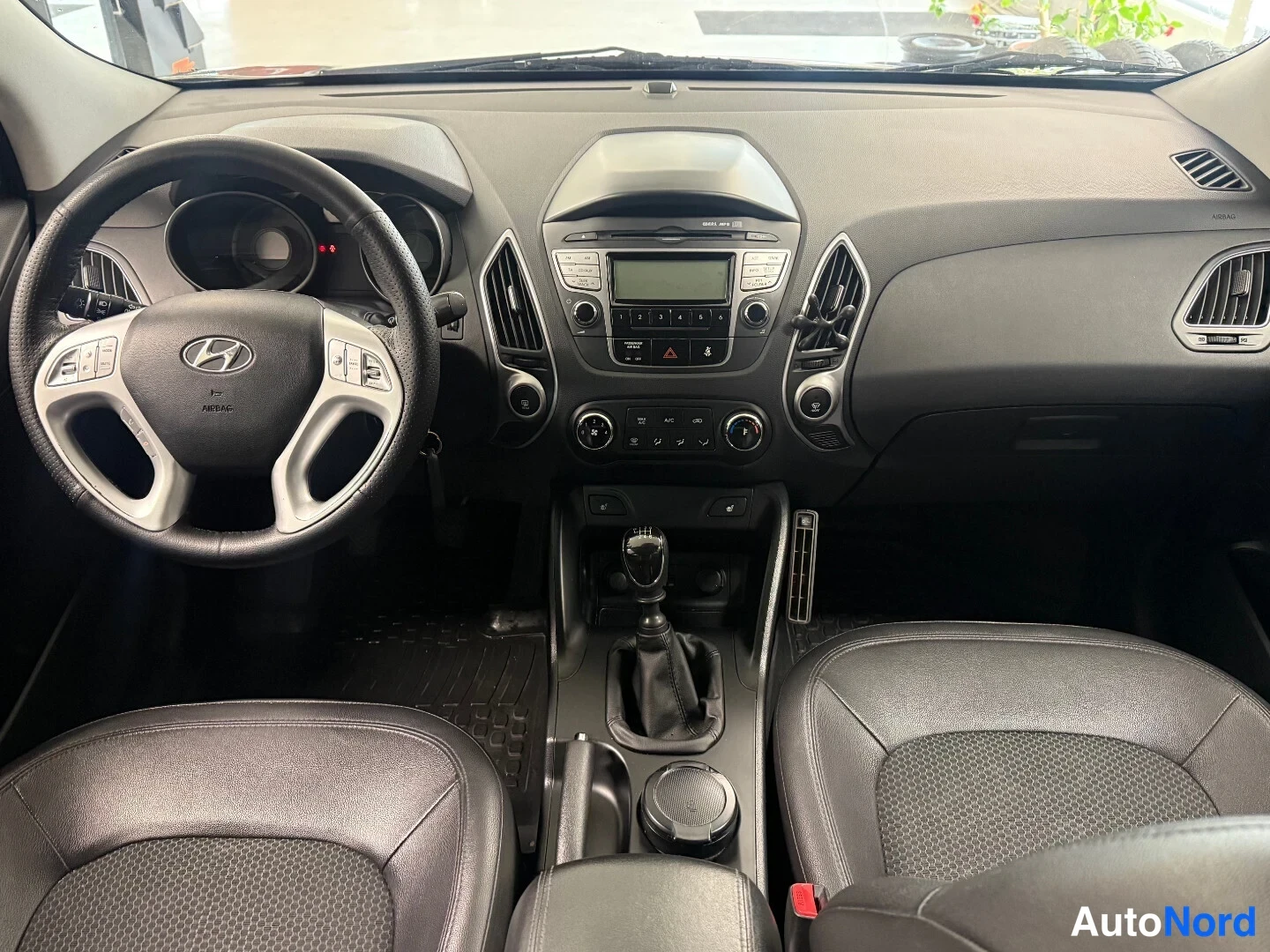 Muu: Hyundai ix35:n ohjaamo suoraan takaa: Hyundai-merkkinen ohjauspyörä vasemmalla, keskikonsolissa radio ja manuaalivaihteen vaihtopyörä, tummat penkit.