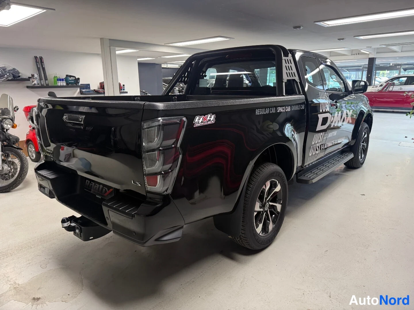 Takaviisto: Mustan avolavan takaa ja oikealta sivulta kuvattu Isuzu D-Max -lainen noutoauto showroomissa; takana vetokoukku ja lavaosassa kaaritanki sekä sivuastin.