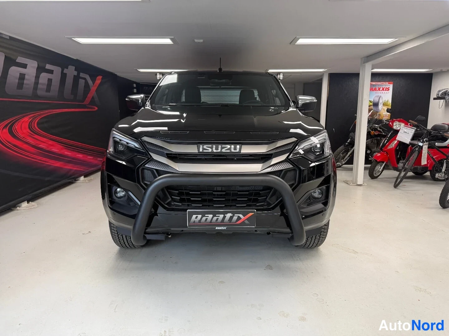 Edestä: Mustan Isuzu D-Max -pakettiauton edustakuva sisätiloissa, ISUZU-merkitty etusäleikkö ja musta suojaus puskurin edessä.
