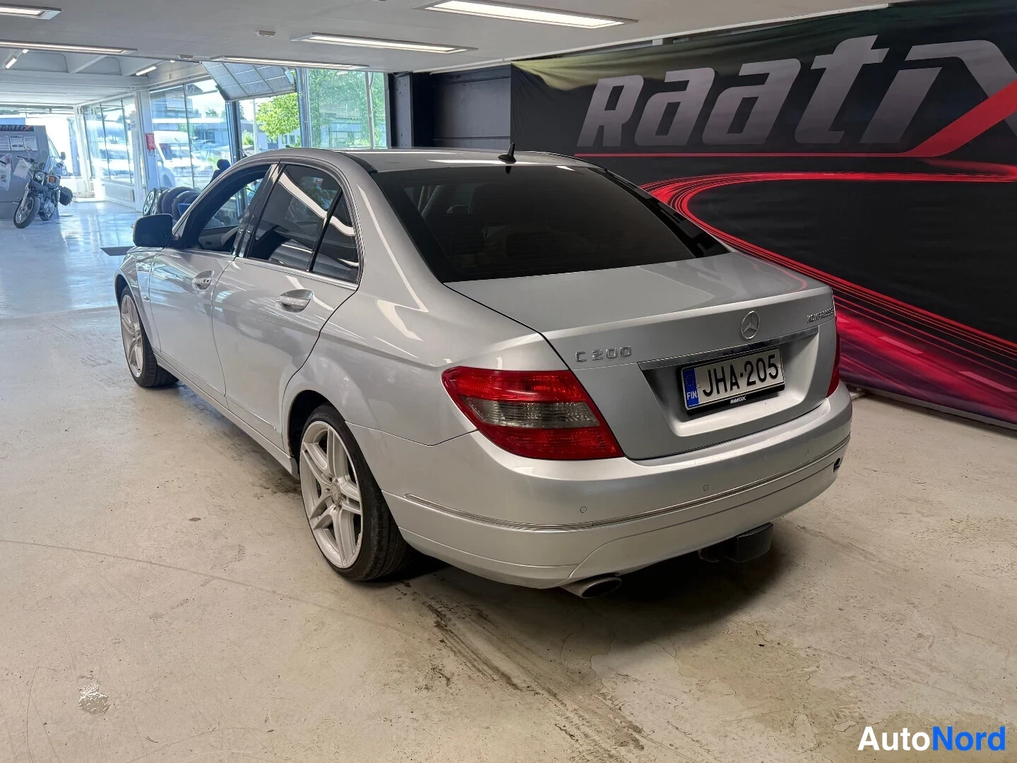 Takaviisto: Hopeisen Mercedes-Benz C 200 -sedanin takaosa ja oikea kylki sisätiloissa, rekisterikilpi.