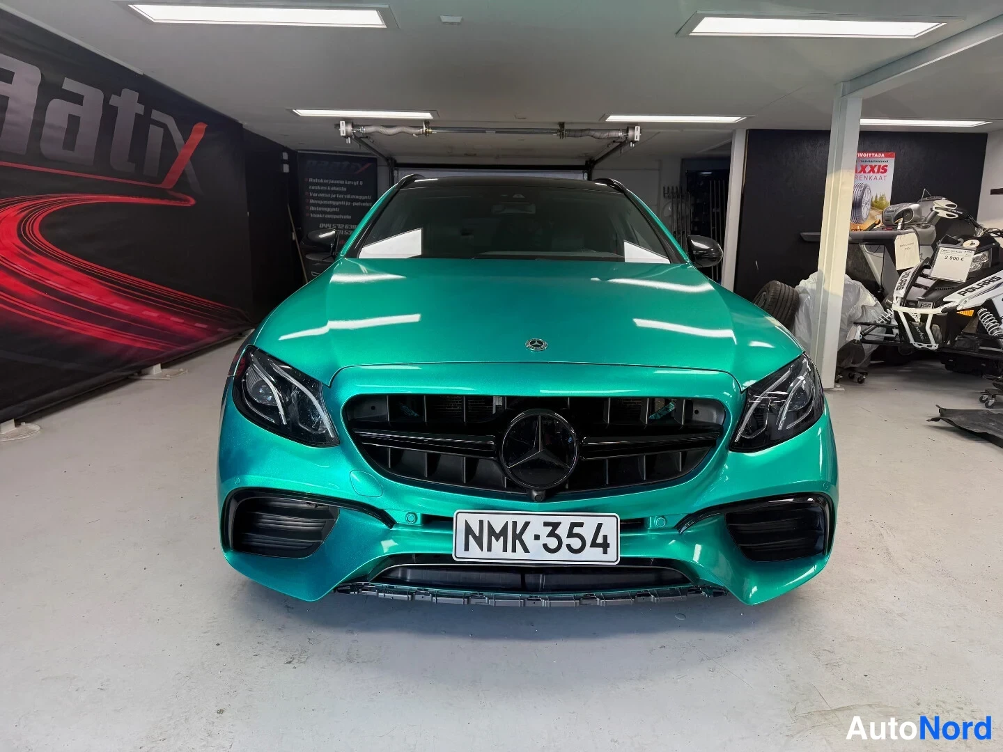 Edestä: Edessäpäin kuvattu Mercedes-Benz E-sarja, tummansinivihreä maali, mustat renovaiset, musta etusäleikkö, rekisterikilpi.