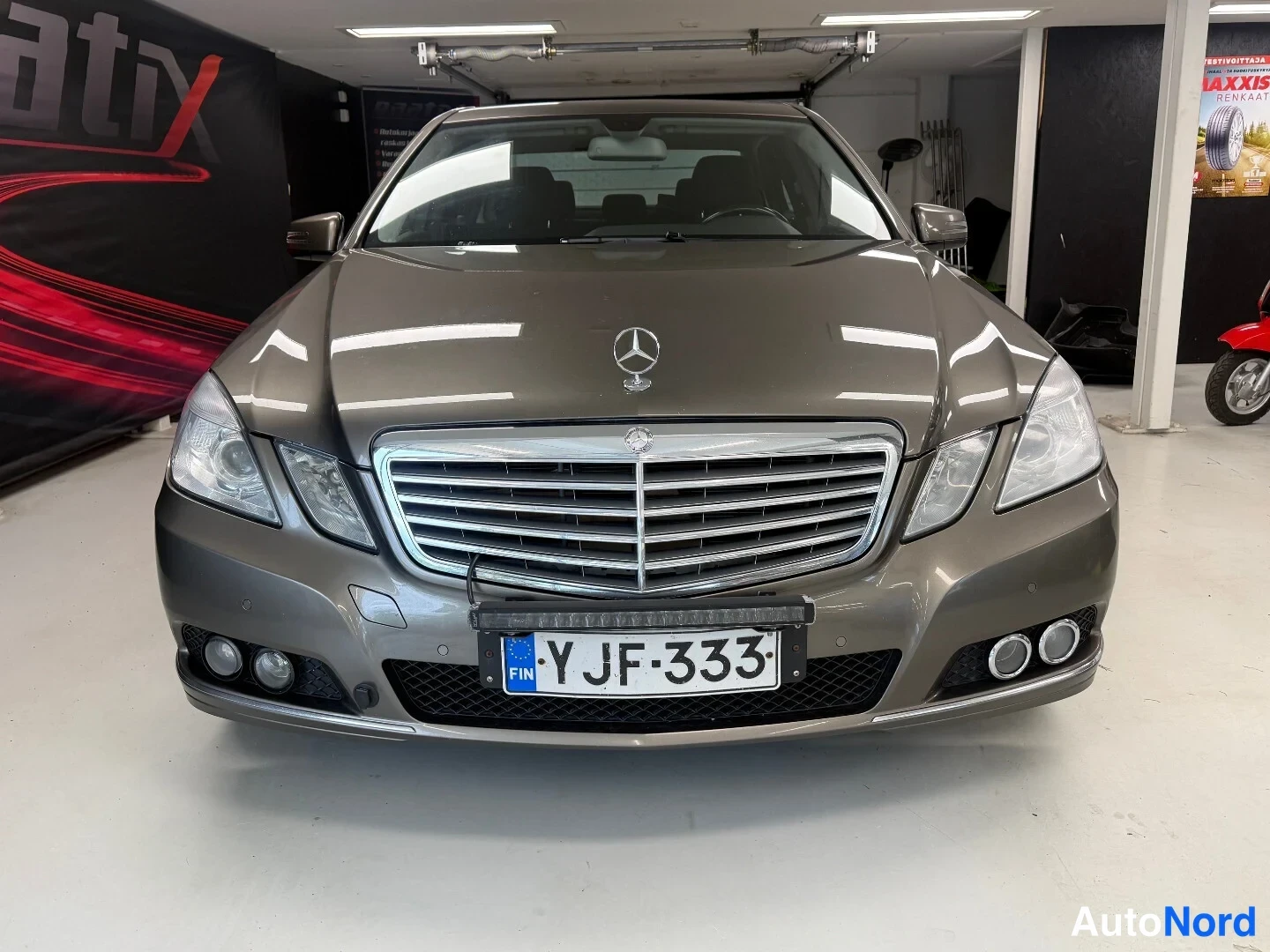Edestä: Mercedes-Benz E-sarjan sedaanin etupääessä katsotaan suoraan eteen; tummanharmaa kori, Mercedes-tähtimerkki konepellillä, rekisterikilpi ja lisävalo]^palkki etu