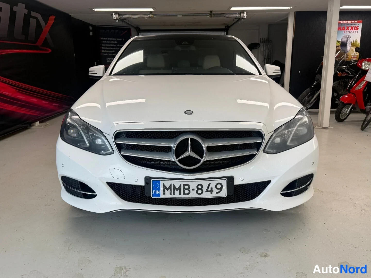 Edestä: Mercedes-Benz E sedanin etupuoli suoraan edestä valokuvattuna; valkoinen kori, Mercedes-tähti etusäleikössä ja EU-kilpi.
