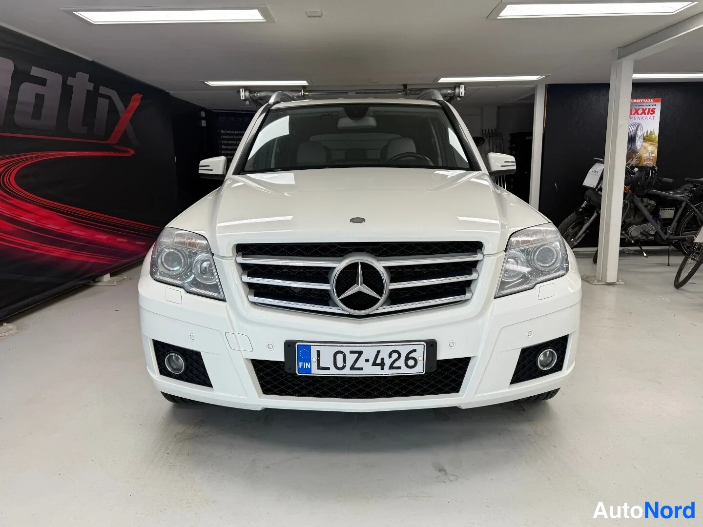 Edestä: Valkoinen Mercedes-Benz GLK -maasturi etukädestä sisätiloissa, rekisteriltään, kattokaiteet asennettuna.