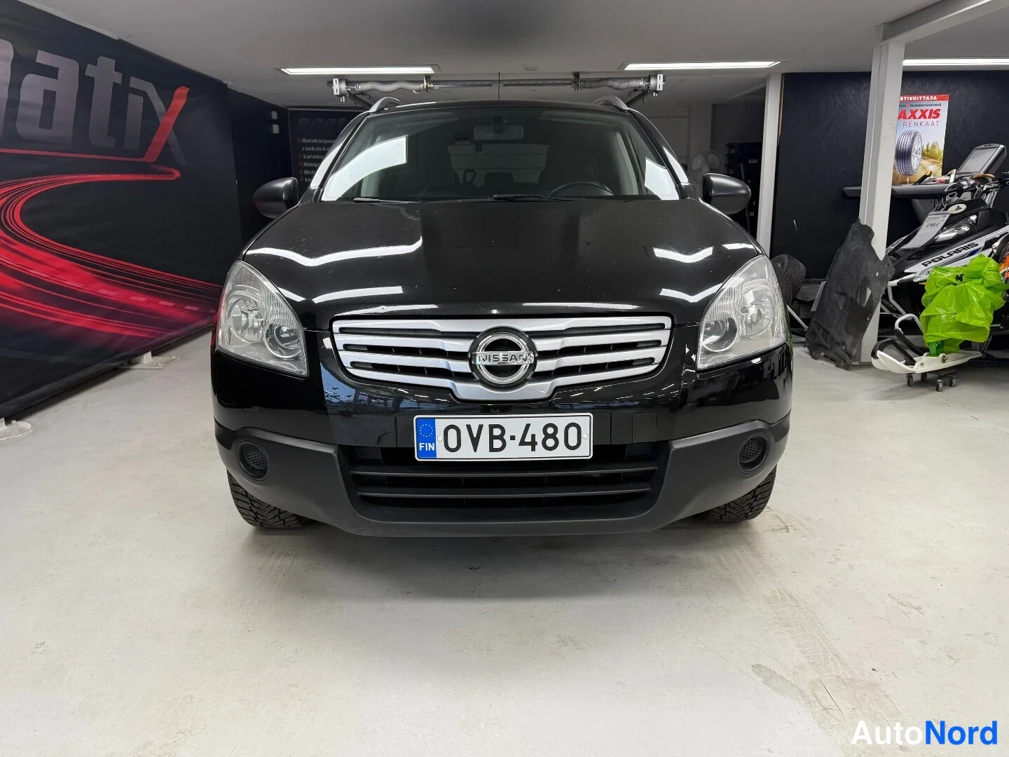 Edestä: Mustan Nissan Qashqai +2 -farmarin etupäät kohtisuoraan edestä kuvattuna; näkyvissä etusäleikkö Nissan-merkillä, ajovalot ja rekisterikilpi sisätiloissa.