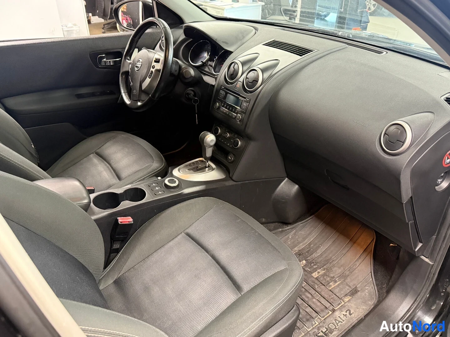 Edestä: Nissan Qashqai +2 -auton ohjaamo vasemmalta ovela kuvattuna: mittaristo, ratit, keskirivin istuimet, automaattivaihde ja lattiamatto.