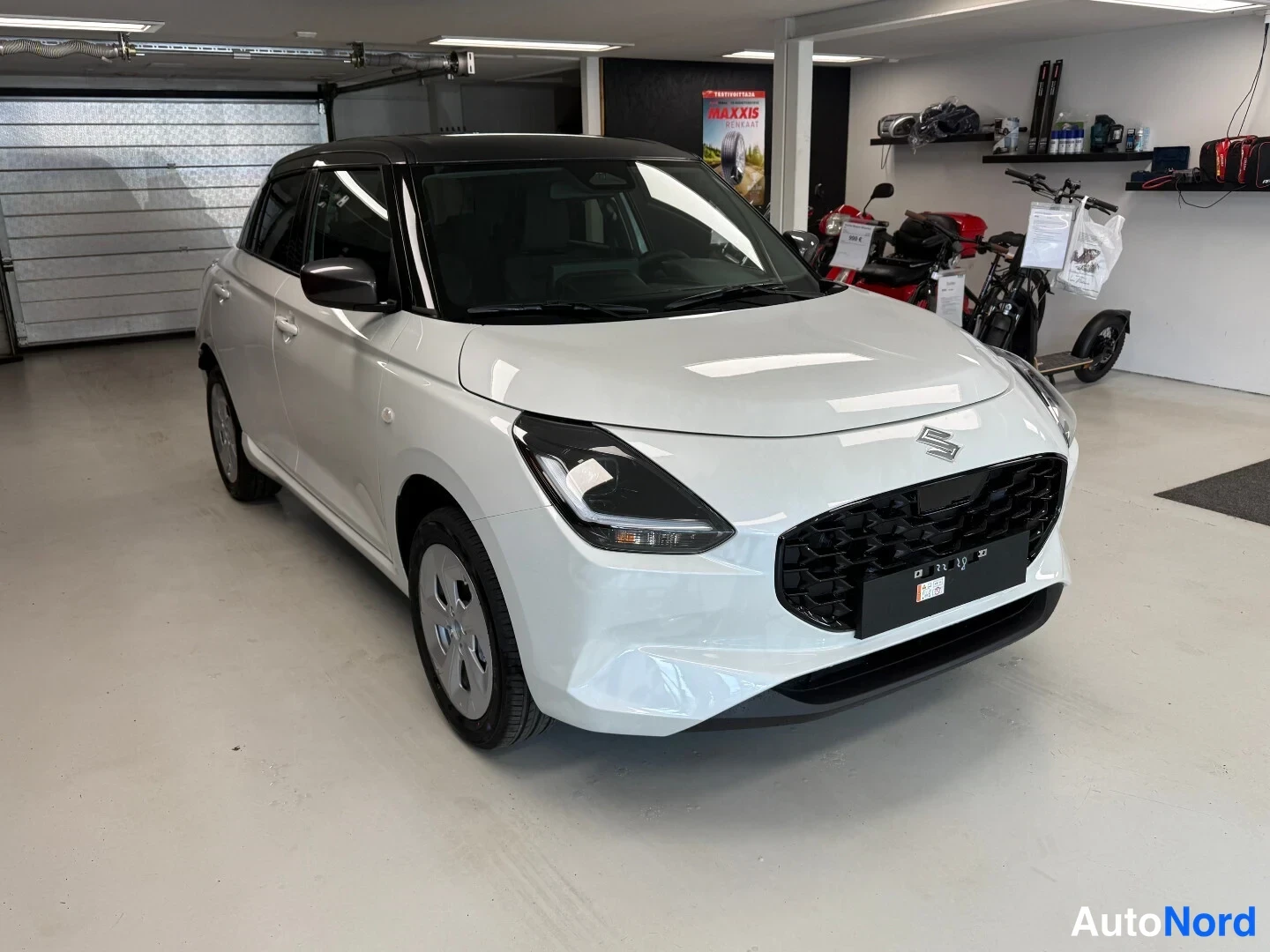 Etuviisto: Valkoinen hatchback edestä ja vasemmalta sivulta kuvattuna autotallissa: musta katto, tumma etusäleikkö ja Suzuki-merkki konepellillä.