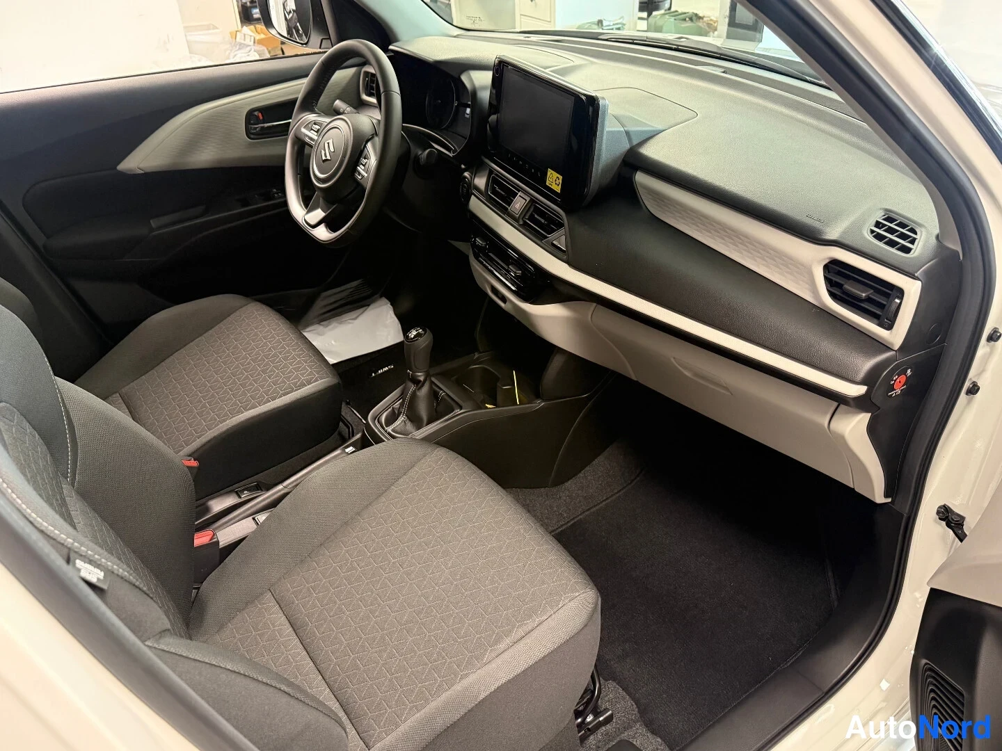 Edestä: Suzuki Swiftin sisätilat: ohjaamo tummilla kankaanistuimilla, manuaalivaihteisto, ohjauspyörä ja kojelaudan infotainment-näyttö.