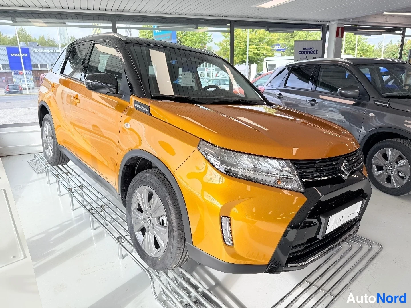 Etuviisto: Oranssi Suzuki Vitara etuosa ja oikea kylki näkyvät autonäyttelyssä muiden autojen välissä.
