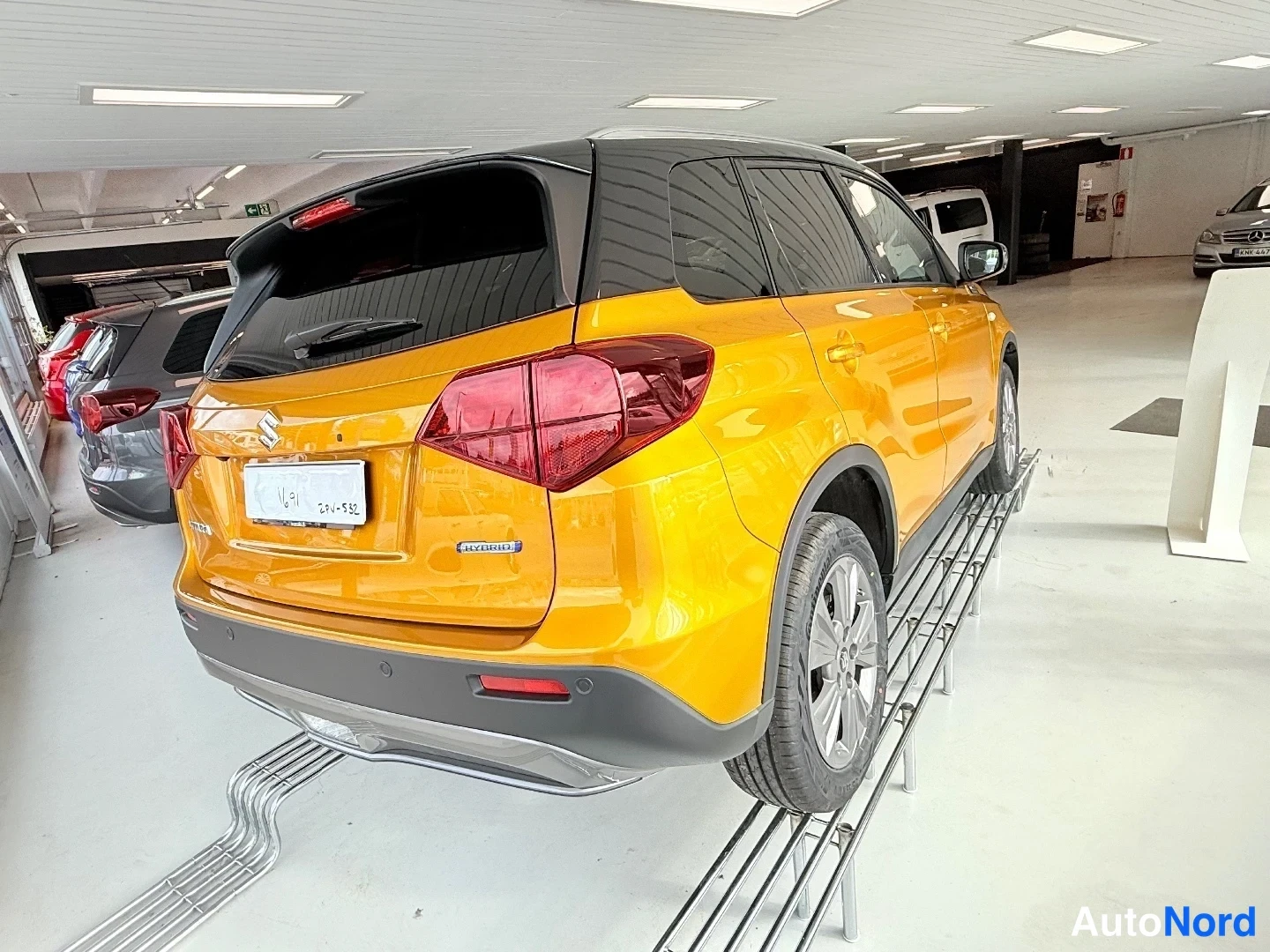 Takaviisto: Oranssi Suzuki Vitara takaa ja vasemmasta kyljestä; peräosassa HYBRID-merkintä, auto näytteillä hallissa.