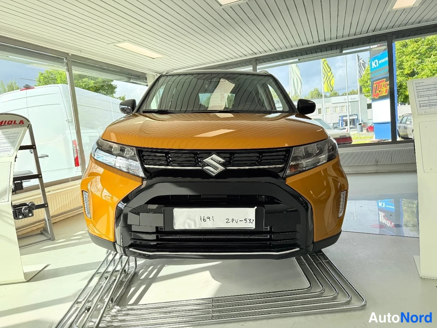 Edestä: Suzuki Vitaran etunäkymä autotilassa; oranssinkeltainen kori, musta etusuoja ja Suzuki-merkki etusäleikössä.