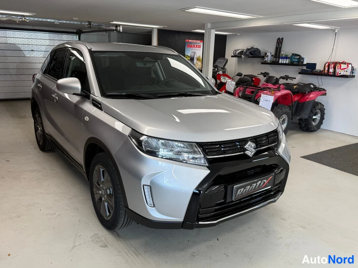 Etuviisto, nokka oikealle: Hopeaharmaa Suzuki Vitara -maastoauto etukulmasta oikeaan viistosti sisätiloissa, keulagrilli ja ajovalot näkyvät.