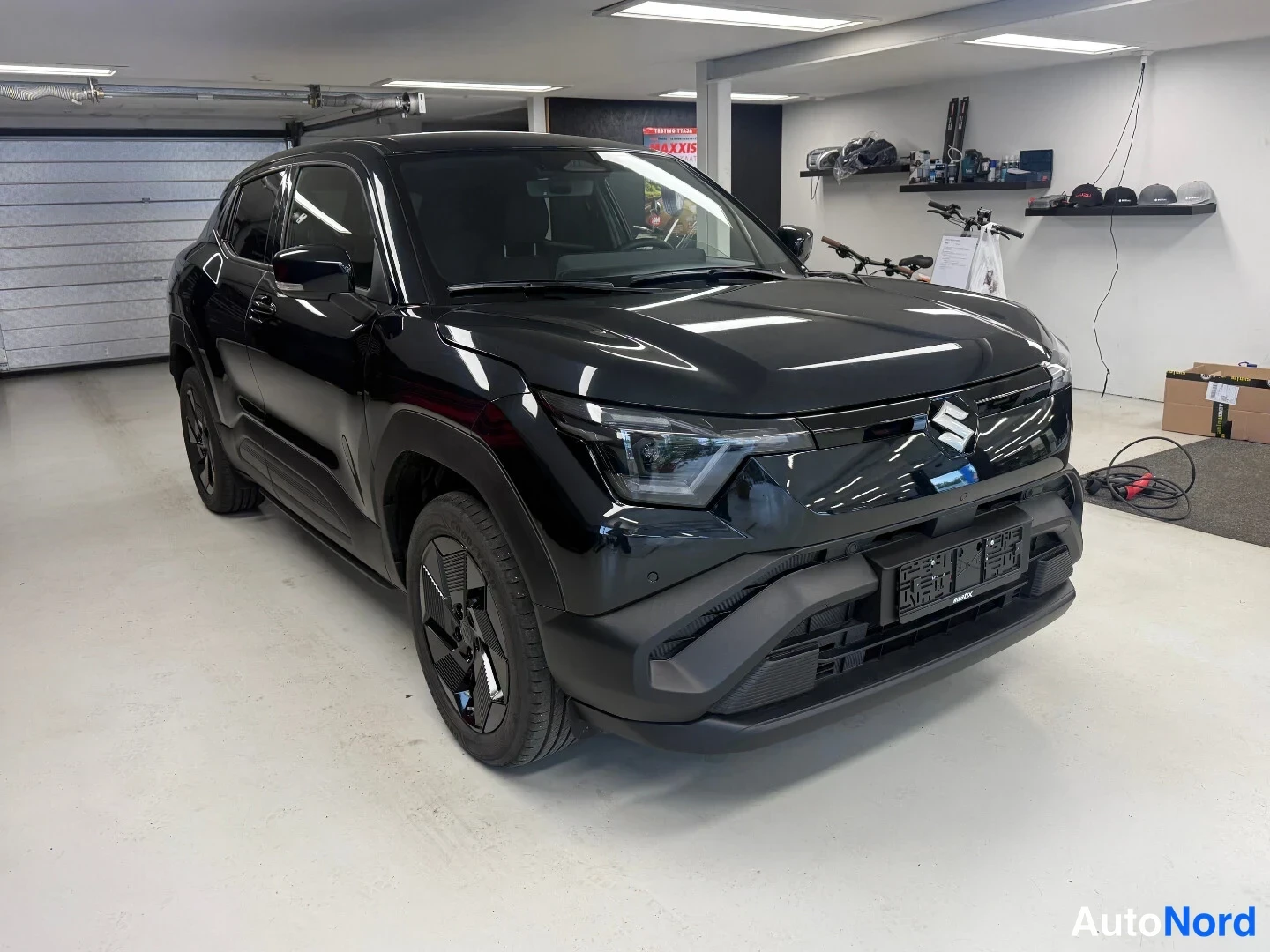 Etuviisto: Mustan Suzuki e-Vitara:n etuosa ja vasen kylki neljännesnäkymässä autotallissa, tyhjä rekisterikilpi edessä.