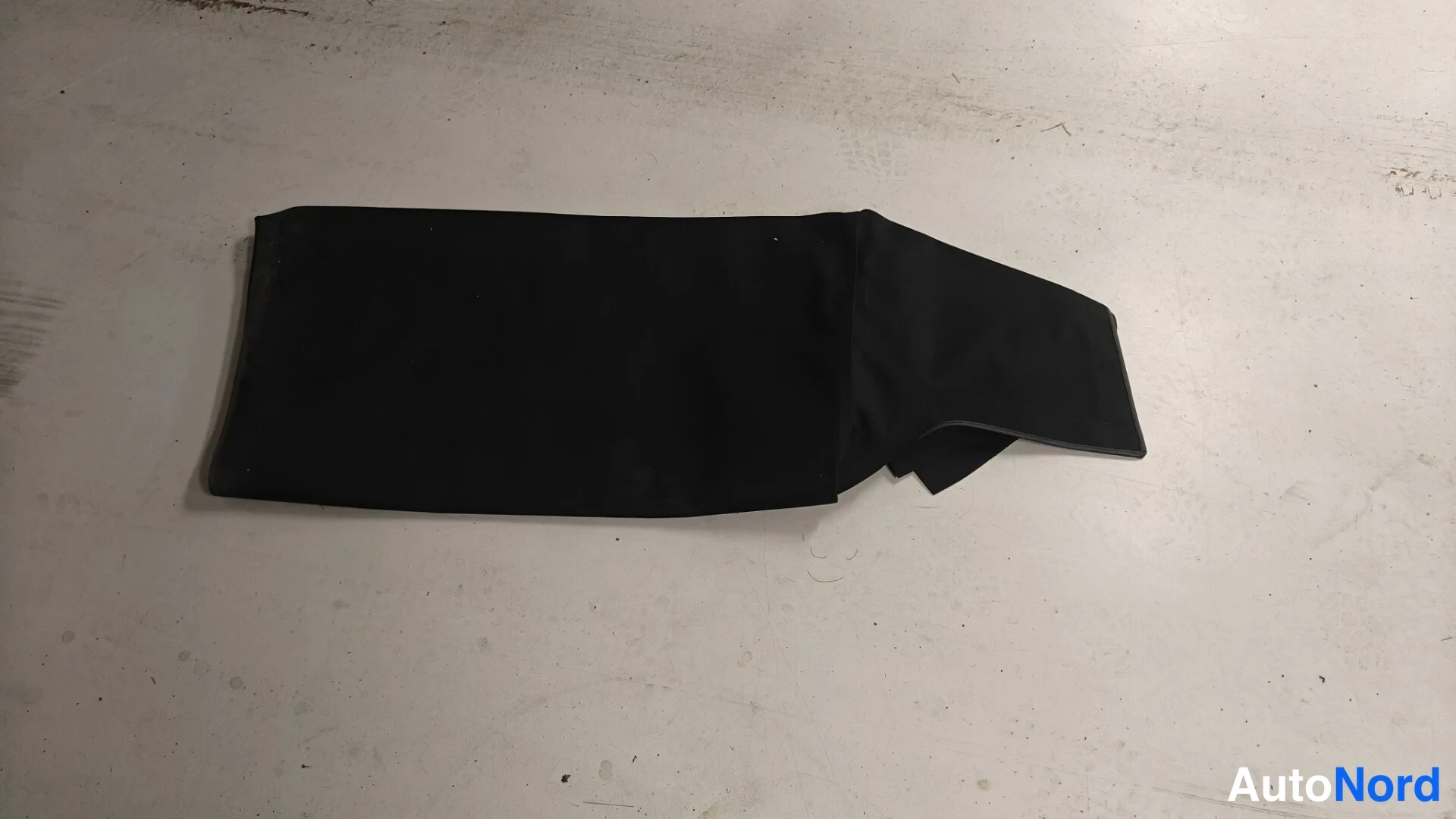 Yksityiskohta: Musta tekstiiliosa, mahdollinen suojapeite tai matto, lattialla vaalealla betonipohjalla.
