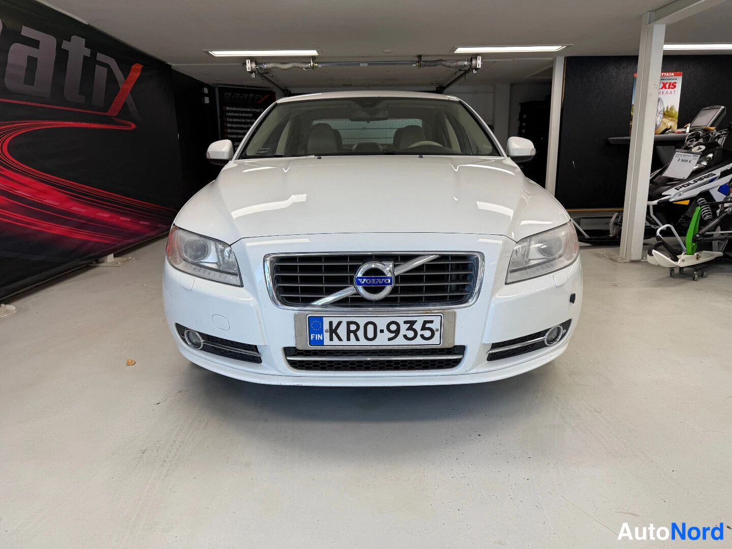Edestä: Valkoisen sedanin etupää, rekisteritunnus, Volvo-merkki etusäleikössä.
