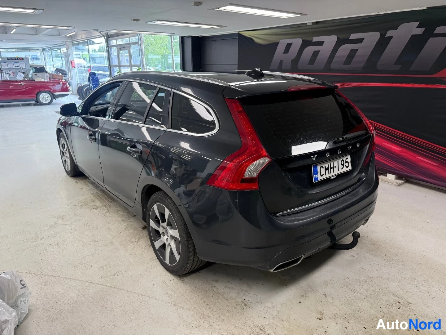 Takaviisto: Tummansininen Volvo V60 -asema-auto takaa oikealta kuvattuna autotallissa; rekisterikilpi ja irtotowbar näkyvät.