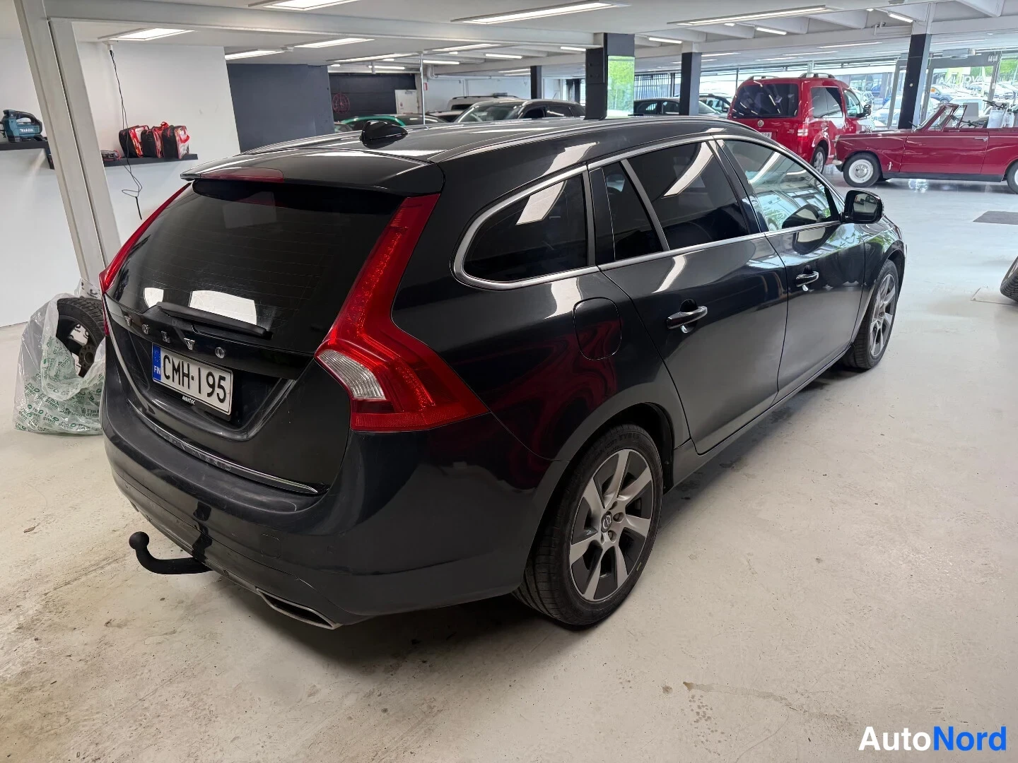 Takaviisto: Tummansininen Volvo V60 -asema-auto takaa vasemmalta kuvattuna autotallissa; rekisterikilpi ja irtotowbar näkyvät.